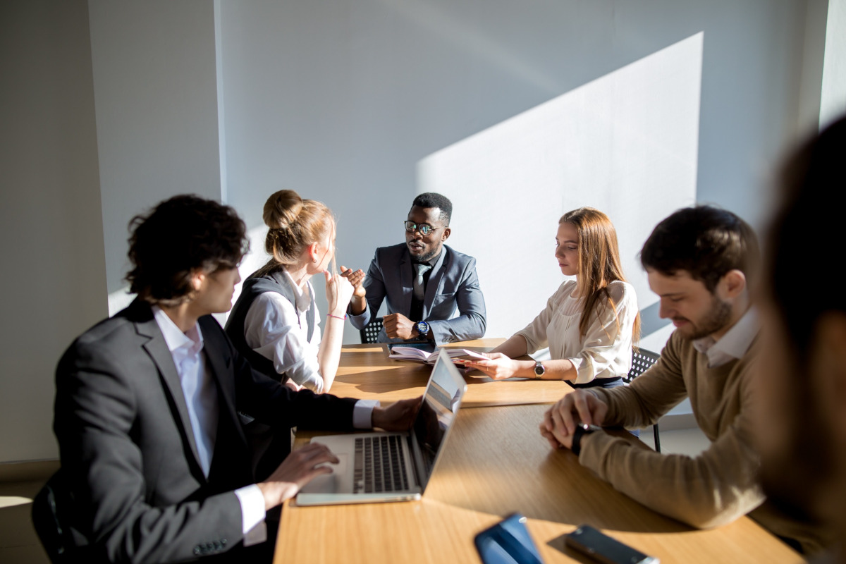A group of business professionals seated around a conference table in a modern office setting, engaged in discussion.